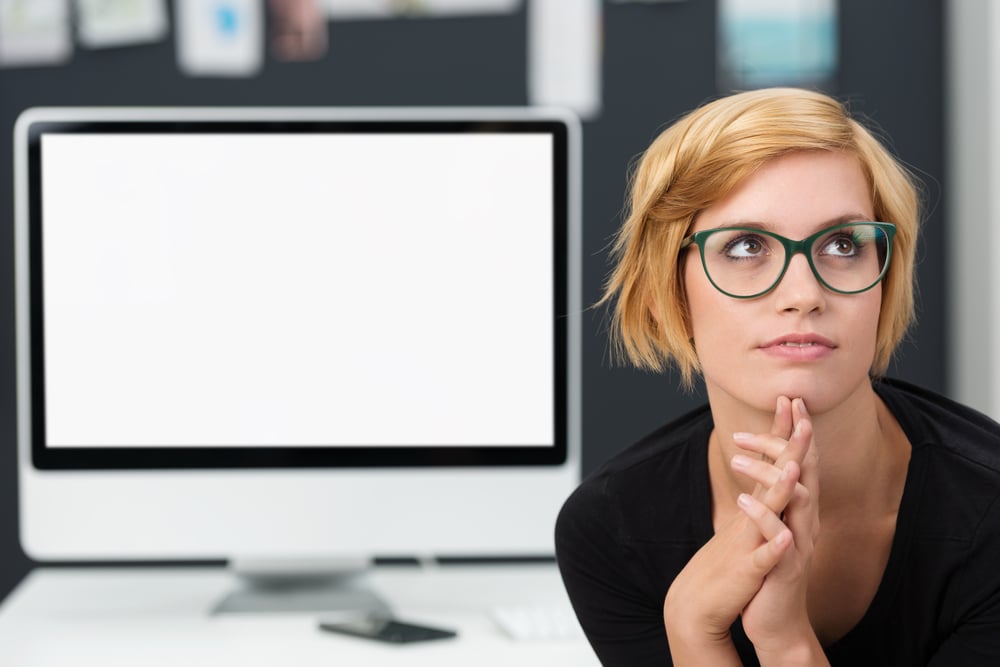 Young businesswoman trying to solve a problem sitting with clasped hands staring thoughtfully into the air in front of a blank desktop monitor with copyspace